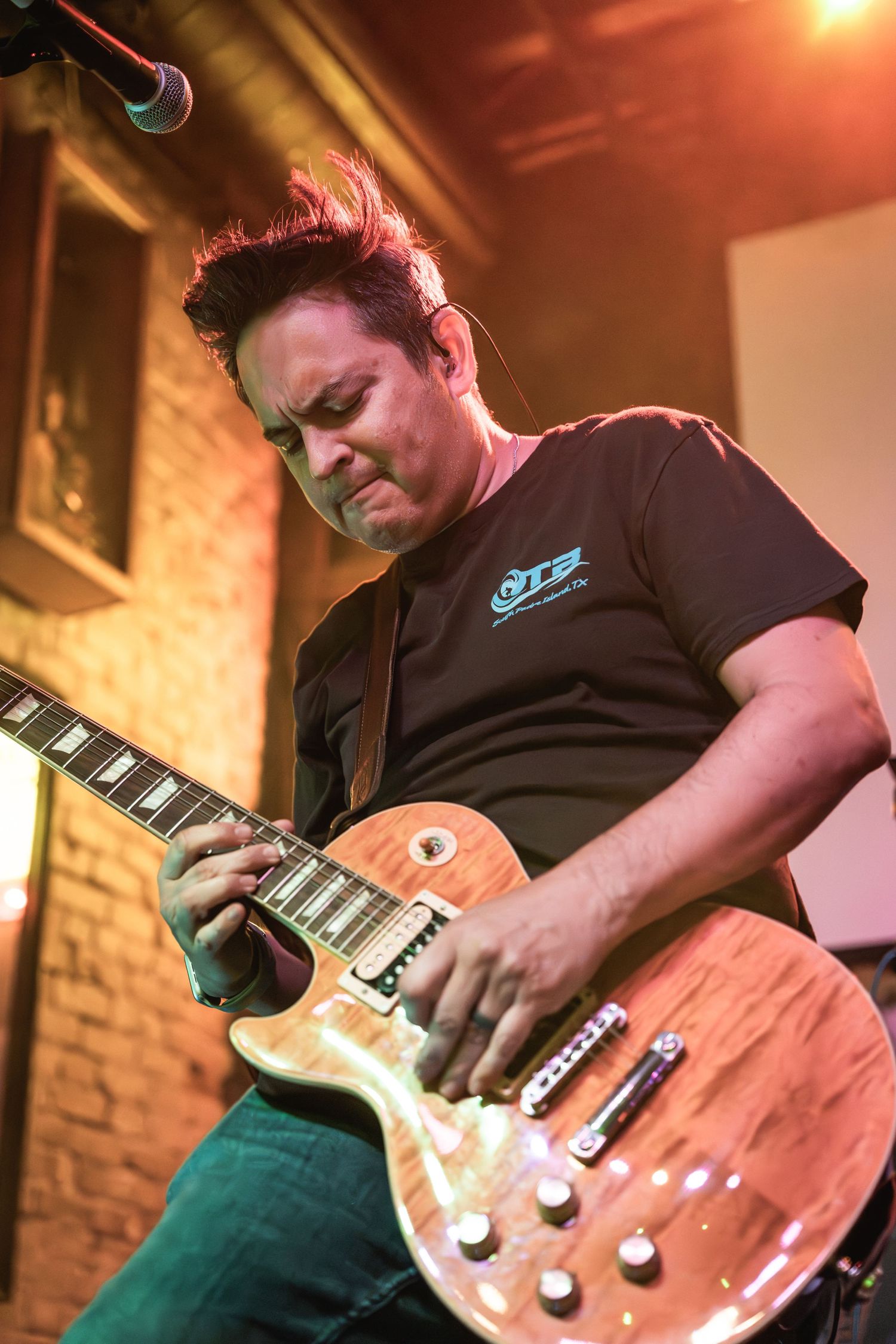 Guitarist in black t-shirt playing Les Paul guitar on stage under dramatic orange lighting.
