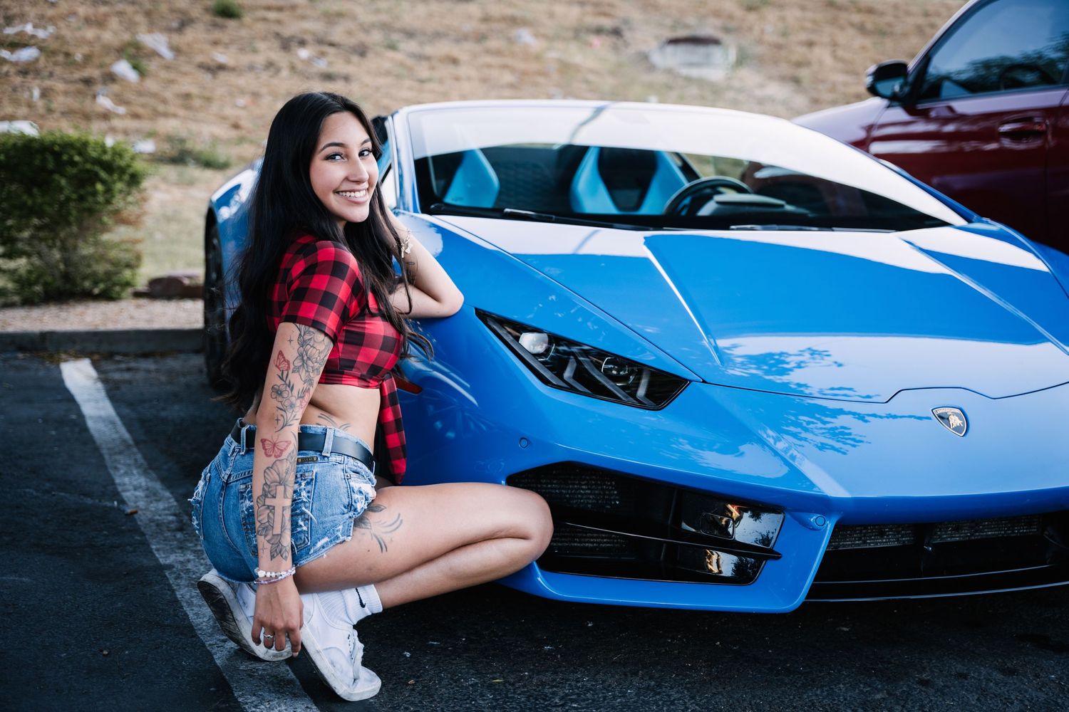 A bright blue Lamborghini photographed with someone in a red plaid top and denim shorts sitting on its front.