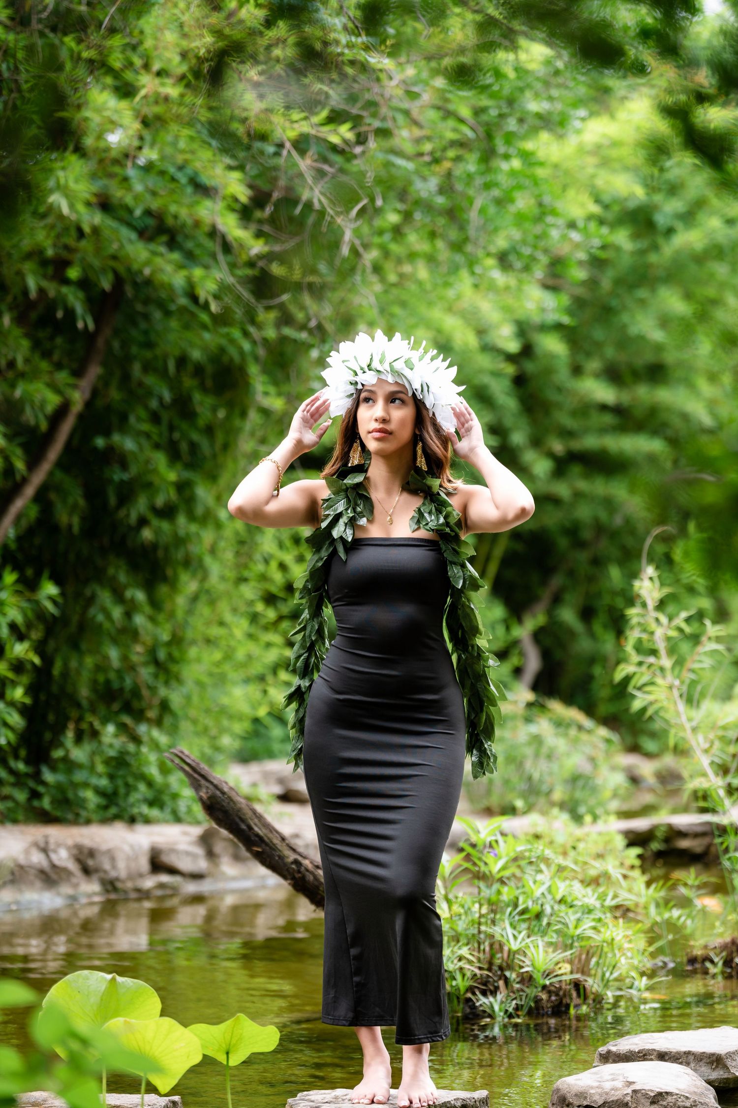 Person wearing black dress and white floral crown poses by water feature in garden setting.