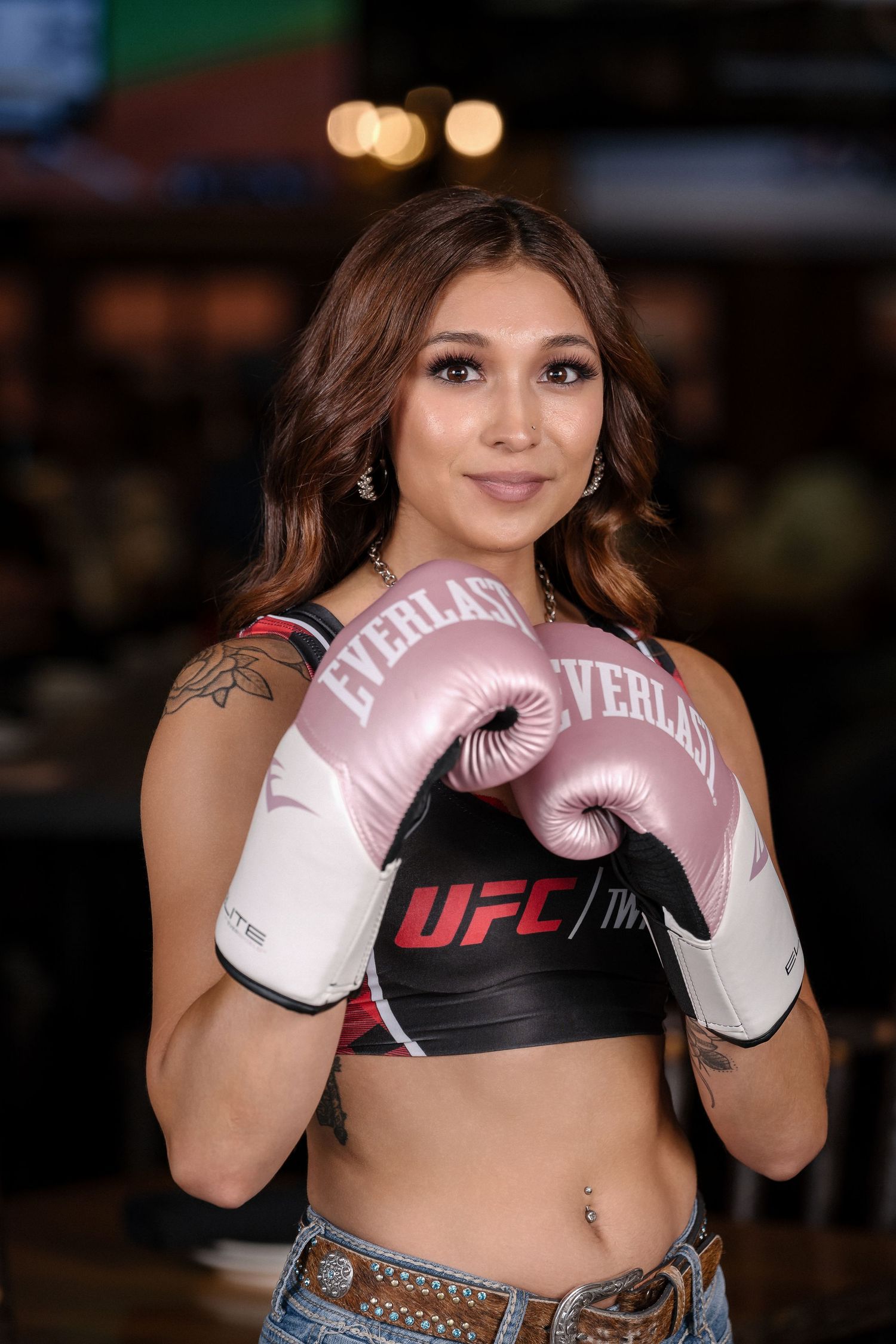 Athletic woman in sports attire poses with pink UFC boxing gloves and crop top in indoor setting.