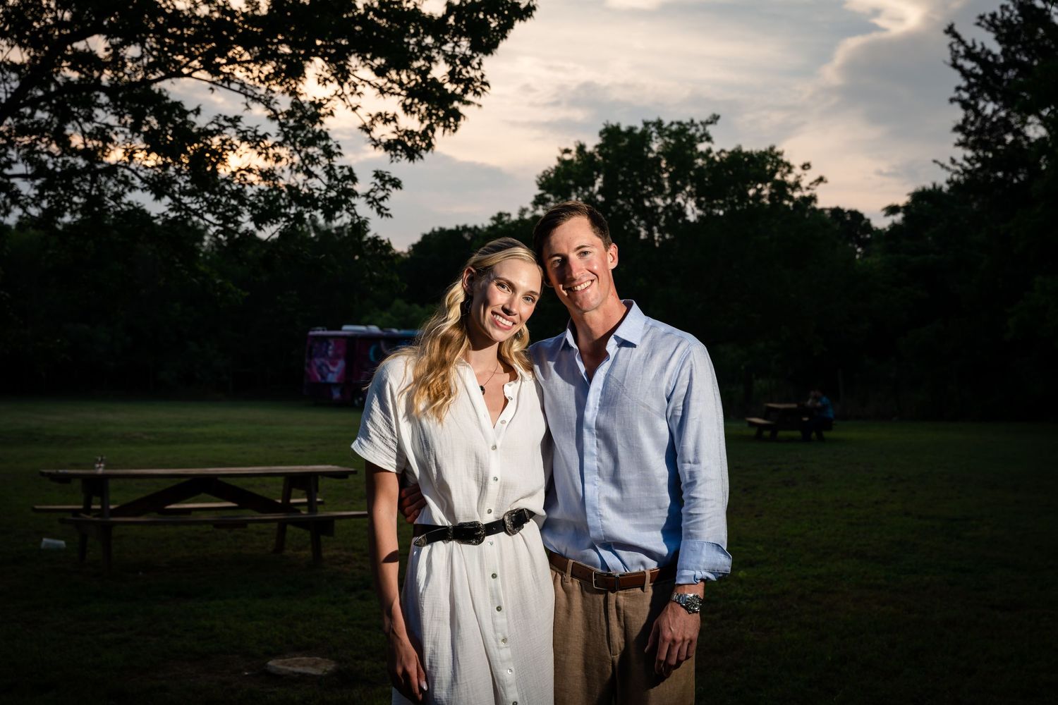 A couple poses together at sunset in a park with trees silhouetted in the background.