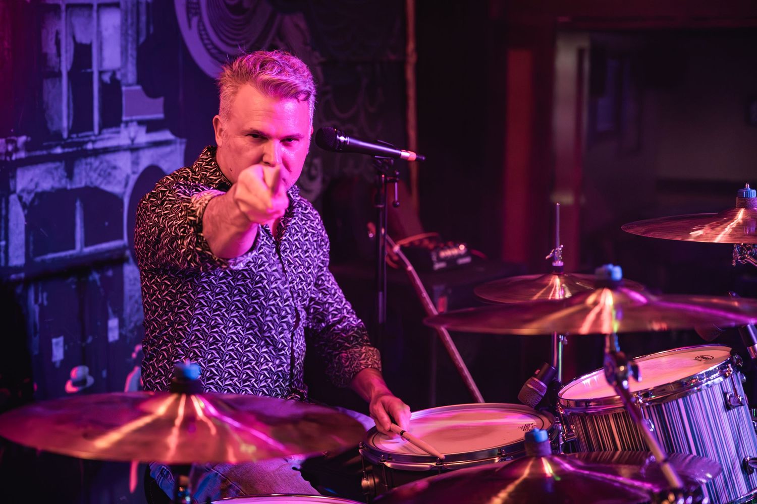 Drummer playing with intense focus under purple stage lights during a live performance.