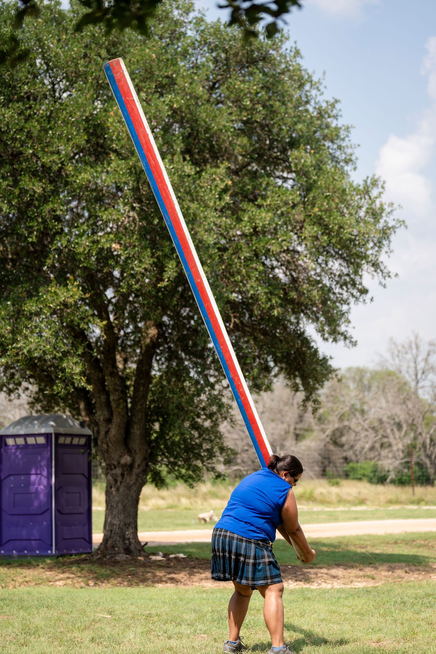 A person in a blue shirt and plaid skirt practices caber toss technique across multiple action shots in a grassy field.