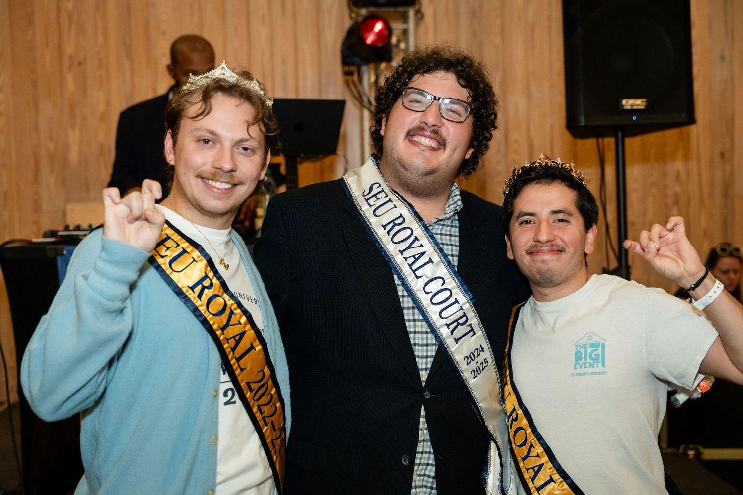 Three students wearing ceremonial sashes pose together at an indoor celebration.