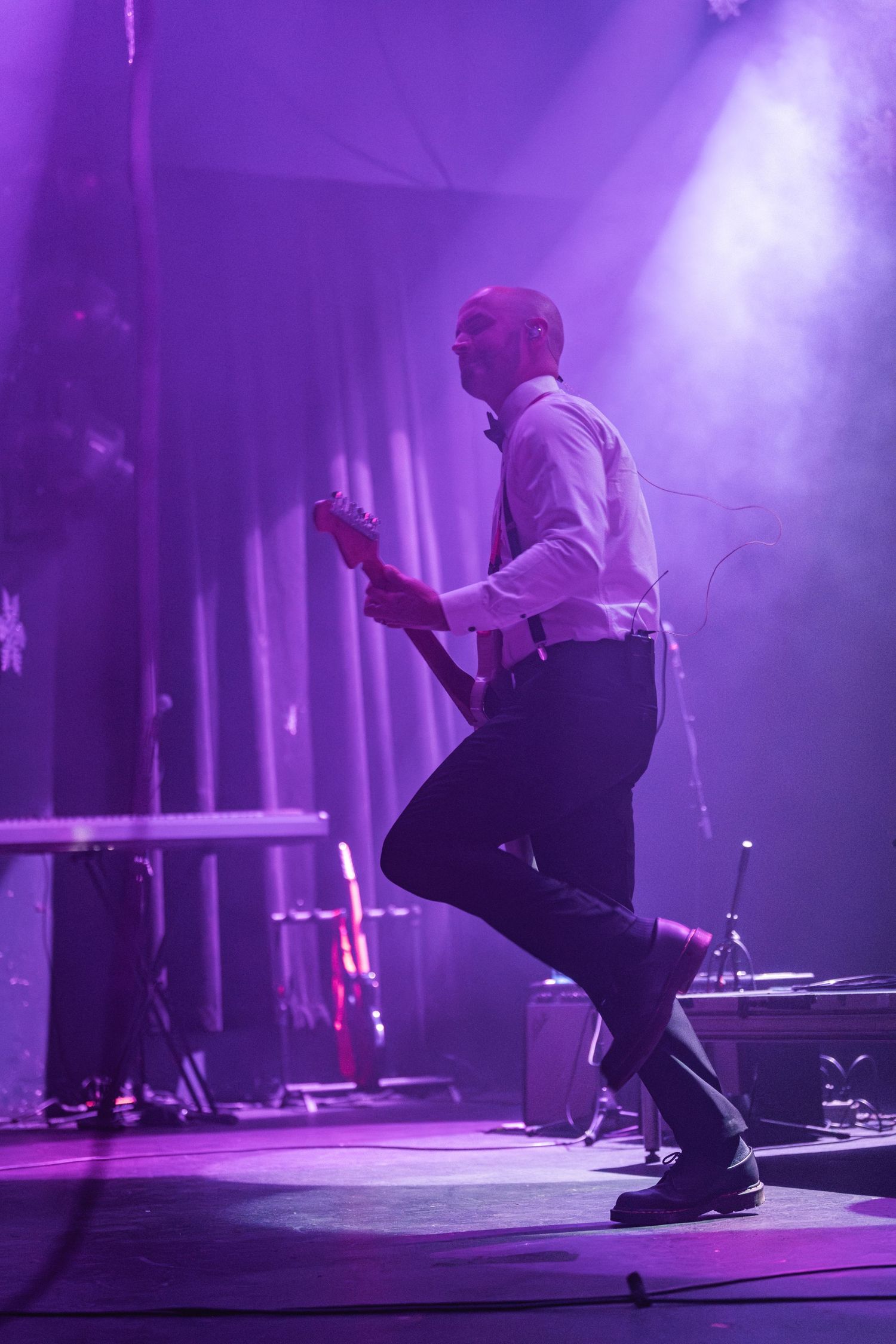 Musician in formal attire plays guitar in dramatic purple stage lighting during performance.