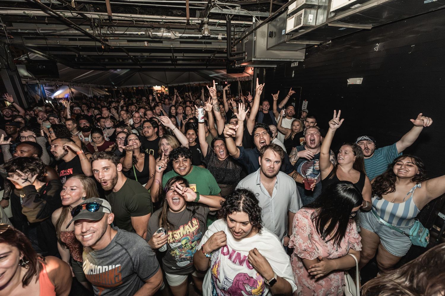 Packed concert venue with enthusiastic crowd raising hands during performance.