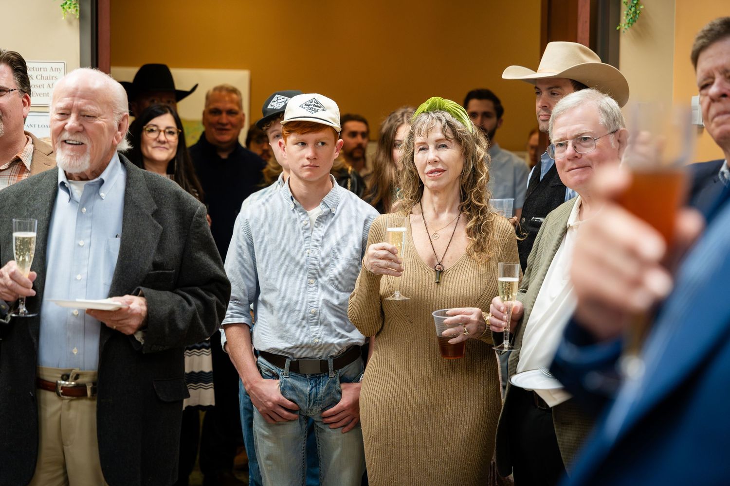 A group of people gather at a social event, with the central figure wearing a tan dress, surrounded by casually dressed guests.