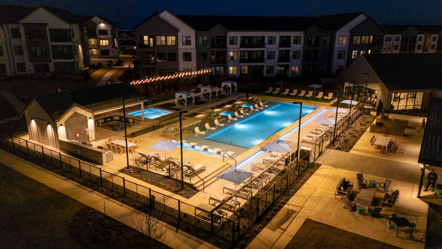 Aerial night view of an illuminated apartment complex swimming pool with lounge chairs and surrounding patio area.