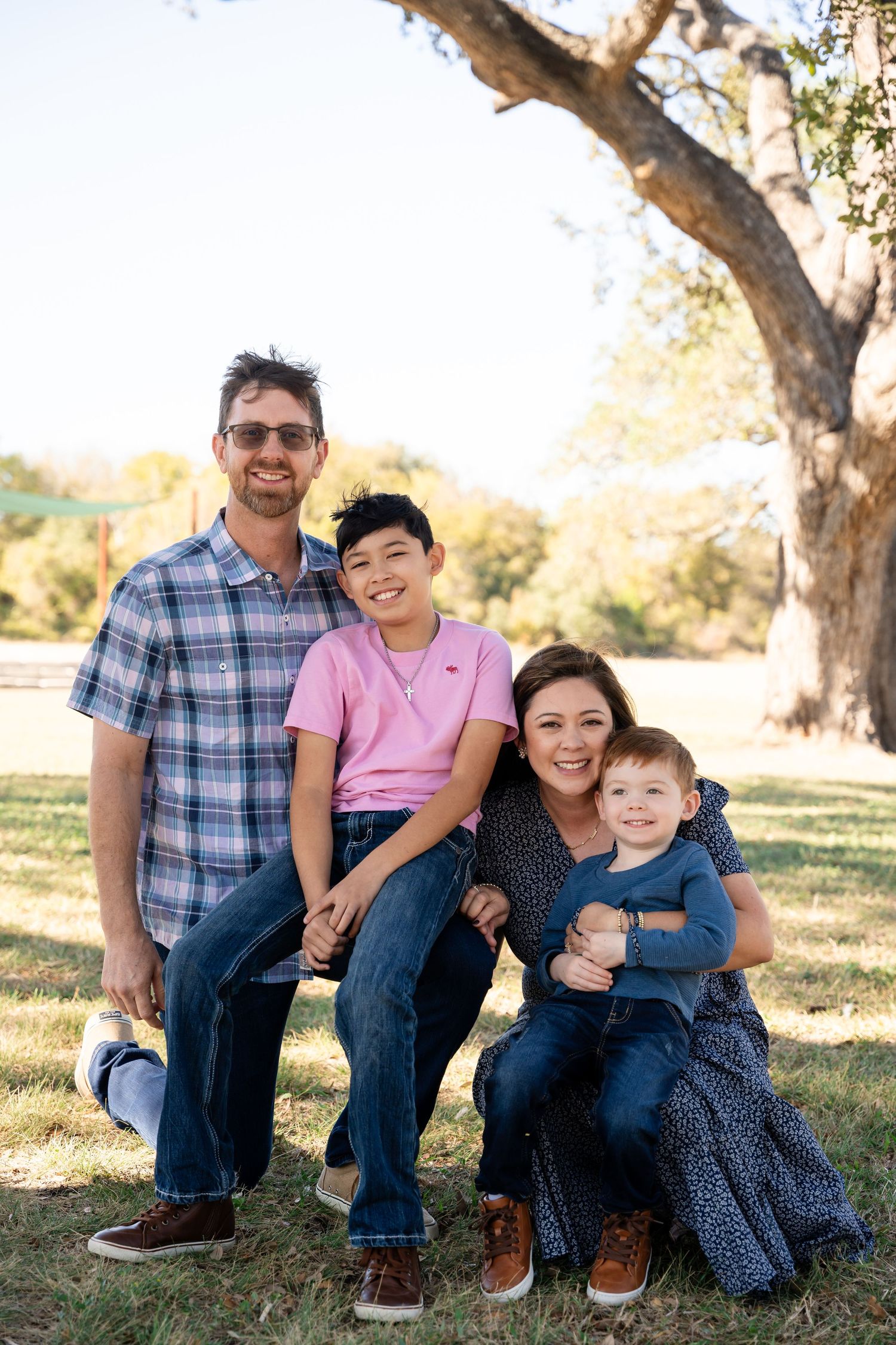 A family sits together on a grassy hill with a large tree in the background.