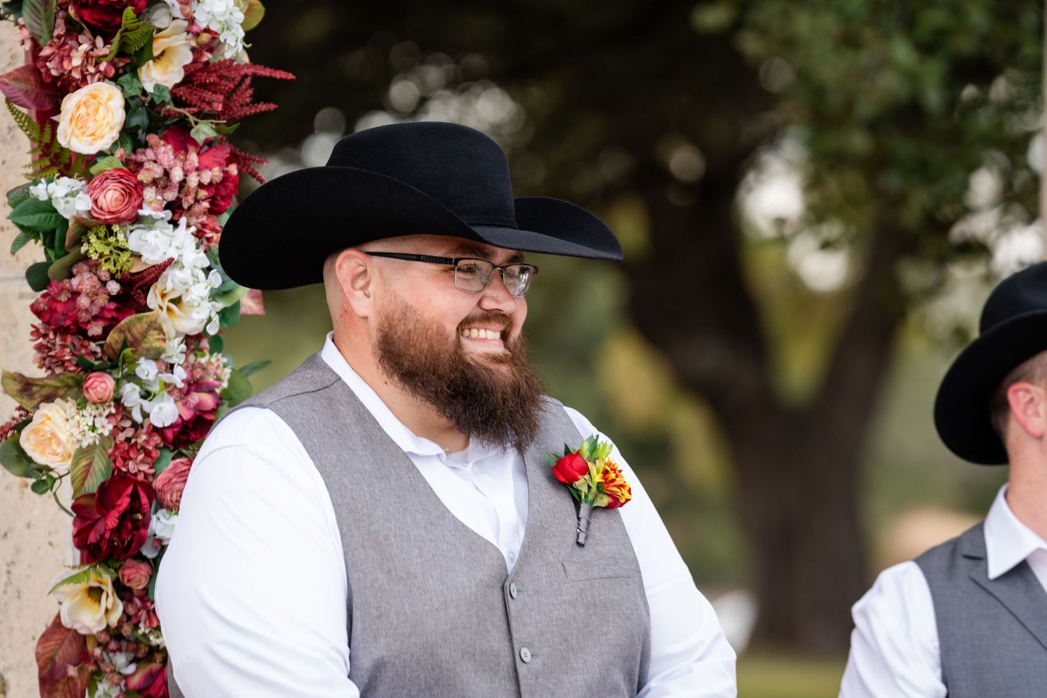 Wedding party member in western attire smiles by floral arch during outdoor ceremony.