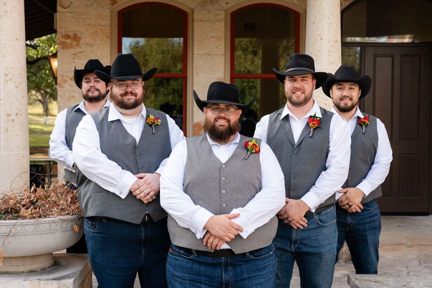 Groomsmen in matching grey vests and cowboy hats standing against stone pillars.