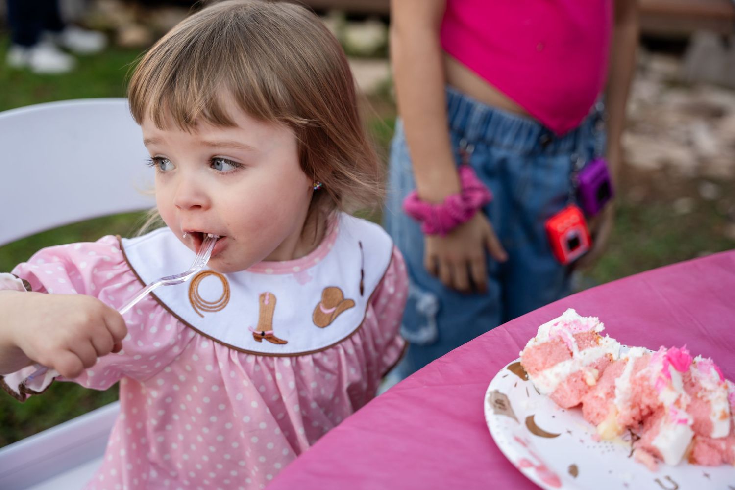 A toddler wearing a polka dot dress sitting at a pink table with birthday cake and watermelon slices.