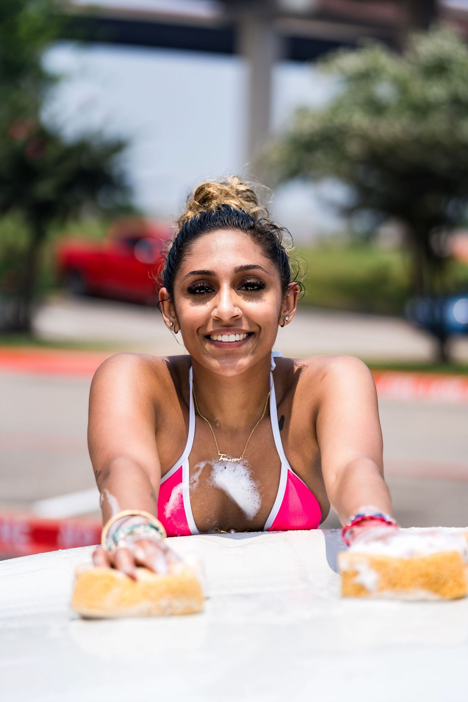 Smiling person in pink swimwear reaching forward during a sunny car wash.