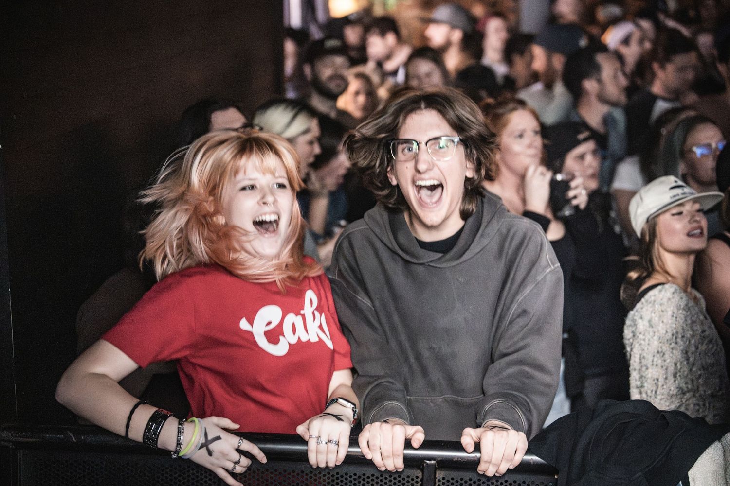 Two excited concert attendees laugh and cheer while leaning on a barrier in a crowded venue.