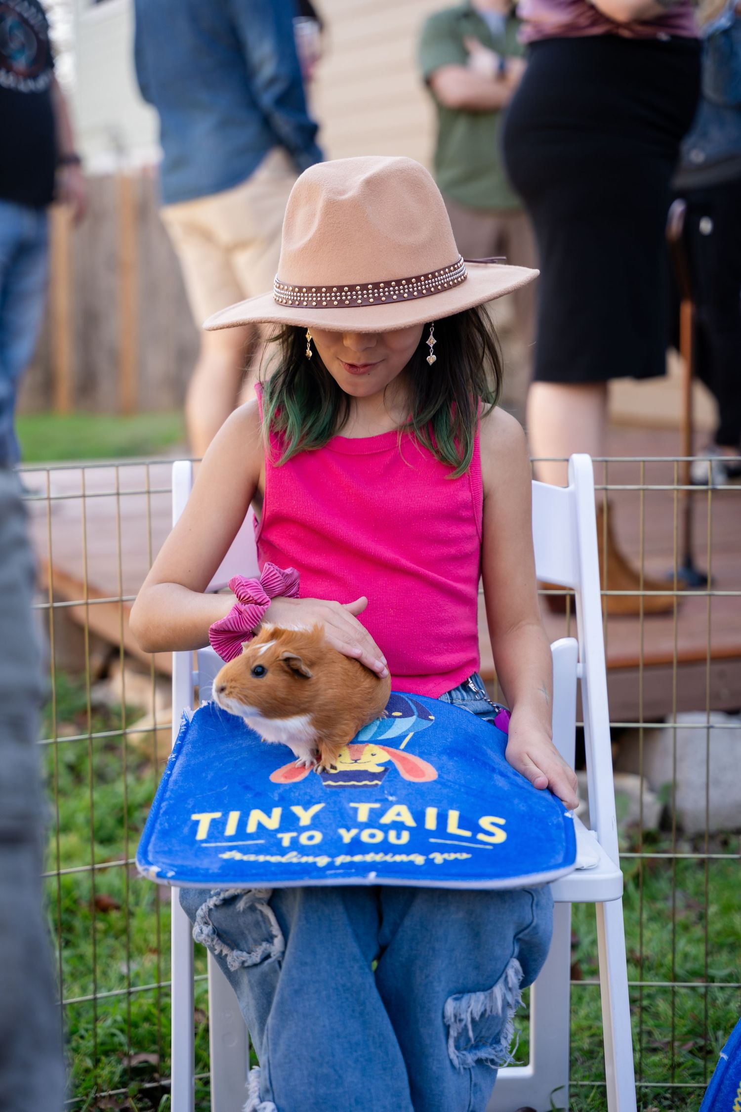 A small guinea pig rests in a Tiny Tails lap while its caregiver wears a pink top and beige hat at an outdoor event.
