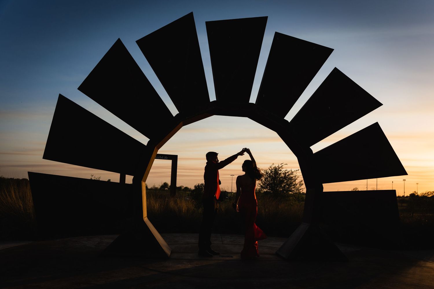 Silhouetted couple dancing within large fan-shaped architectural structure at sunset.