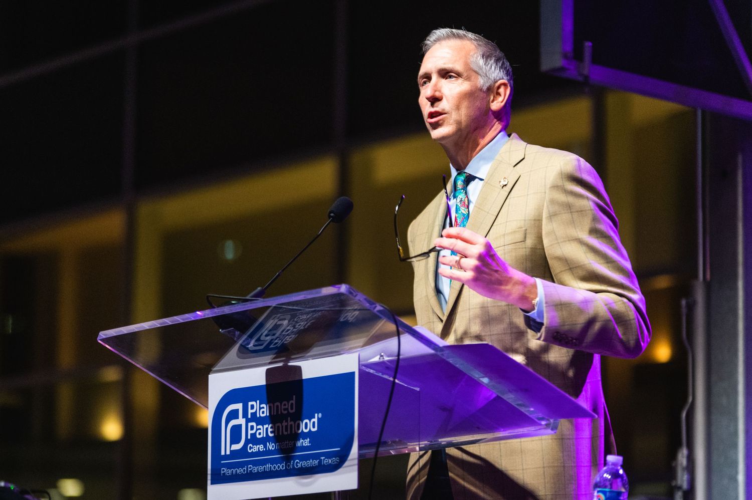 Speaker in beige suit gestures while delivering remarks at branded podium under purple lighting.
