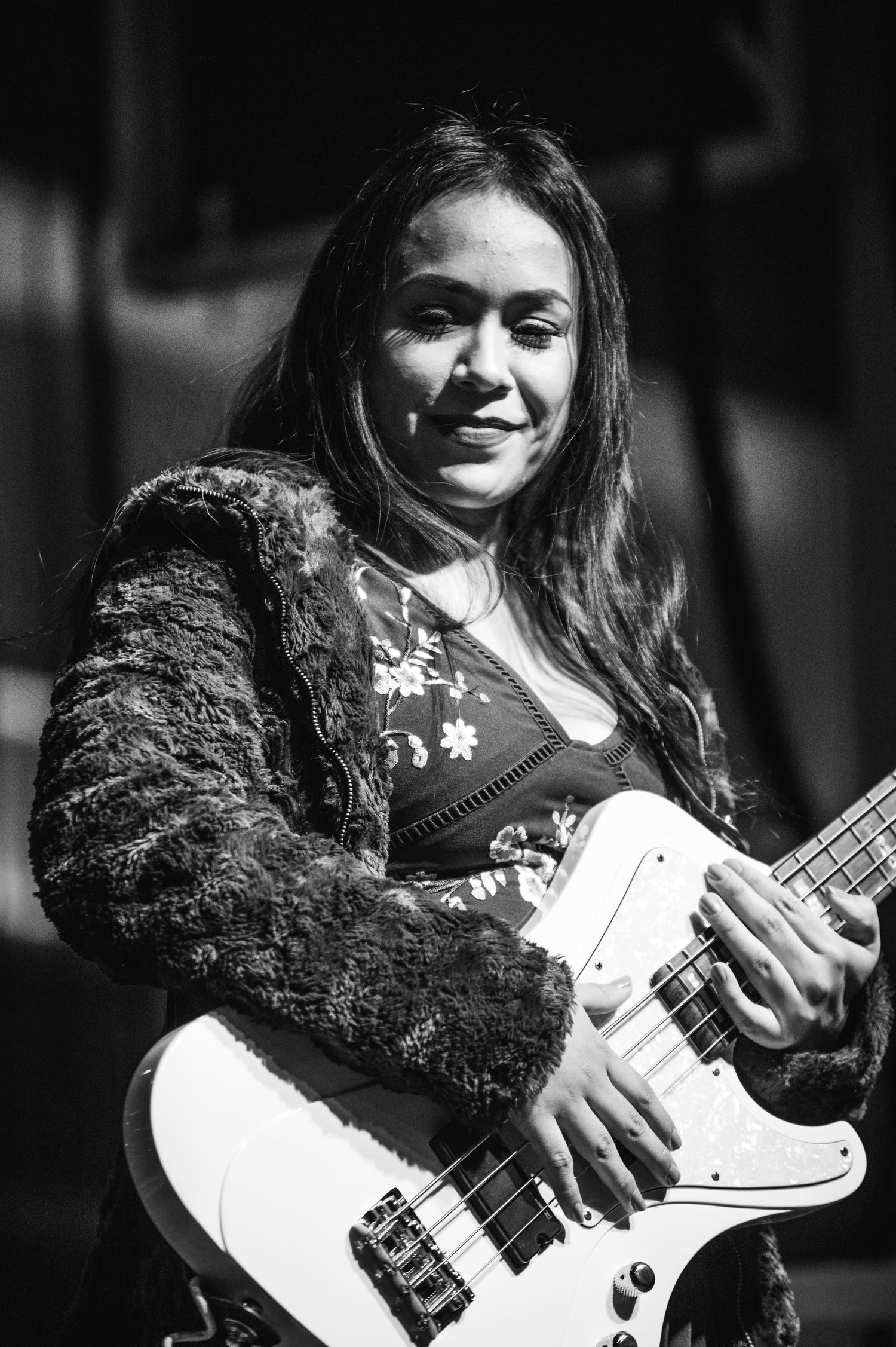 Black and white portrait of bassist performing with white electric guitar and lace jacket on stage.