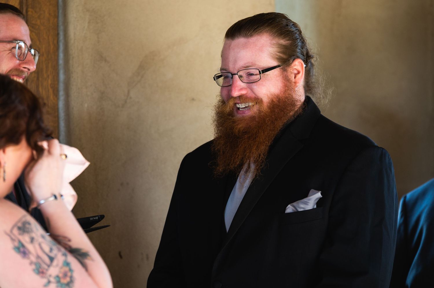A formal wedding participant stands in a black suit with a light pocket square.