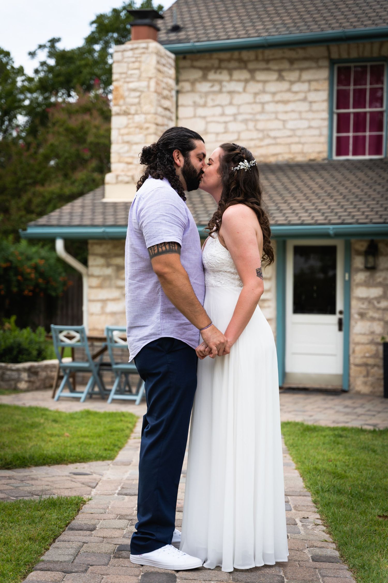 A couple in wedding attire share a romantic moment in front of a stone house.