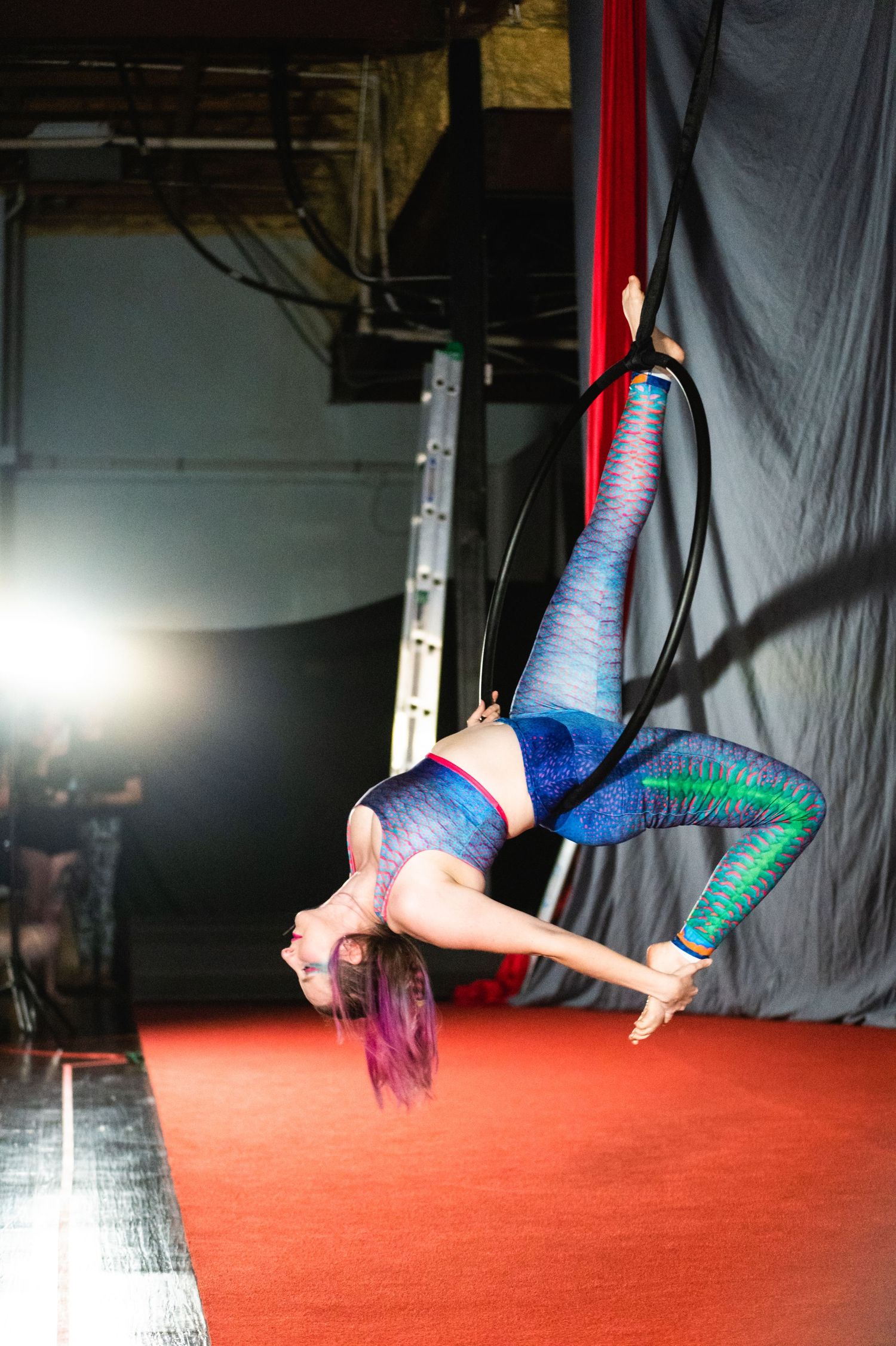 Performer in colorful leggings executes an aerial trick on red silks against dark background.