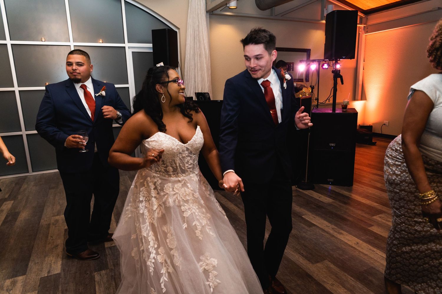 Wedding guests dance and celebrate on a wooden dance floor at an indoor reception venue.