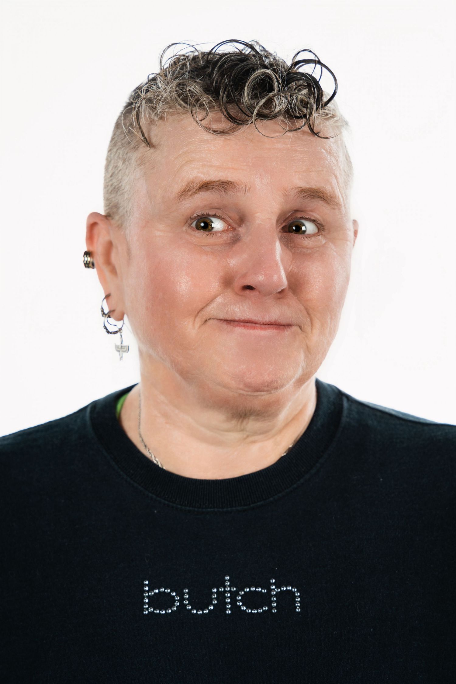 Person in black shirt with curly hair and multiple earrings smiles at camera against white background.