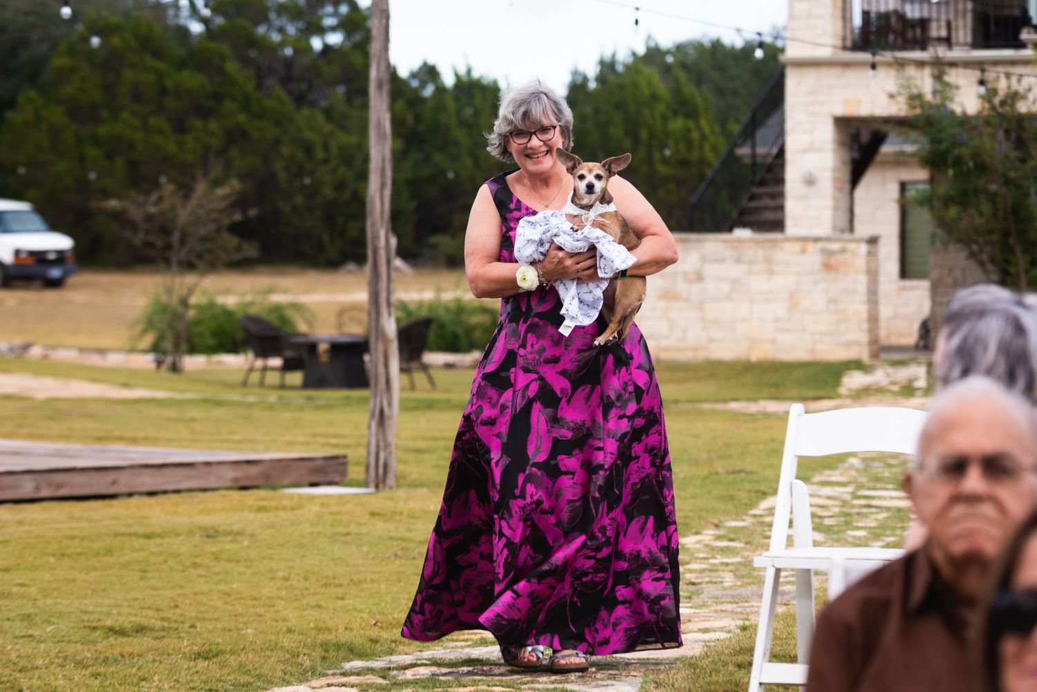 A person in a purple floral dress carries a small dog during an outdoor event.