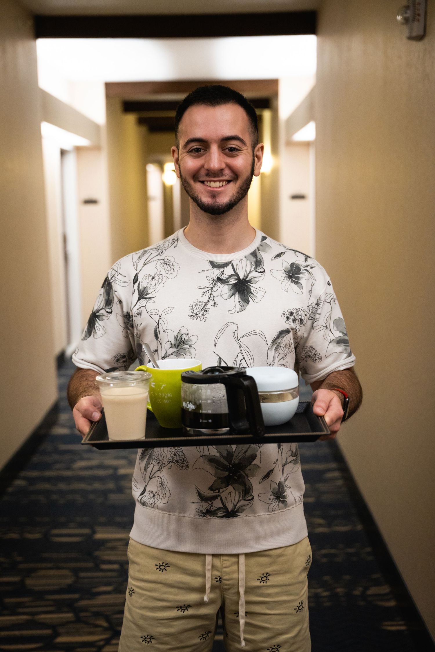 Server carrying tray with drinks in hotel hallway wearing floral print t-shirt.