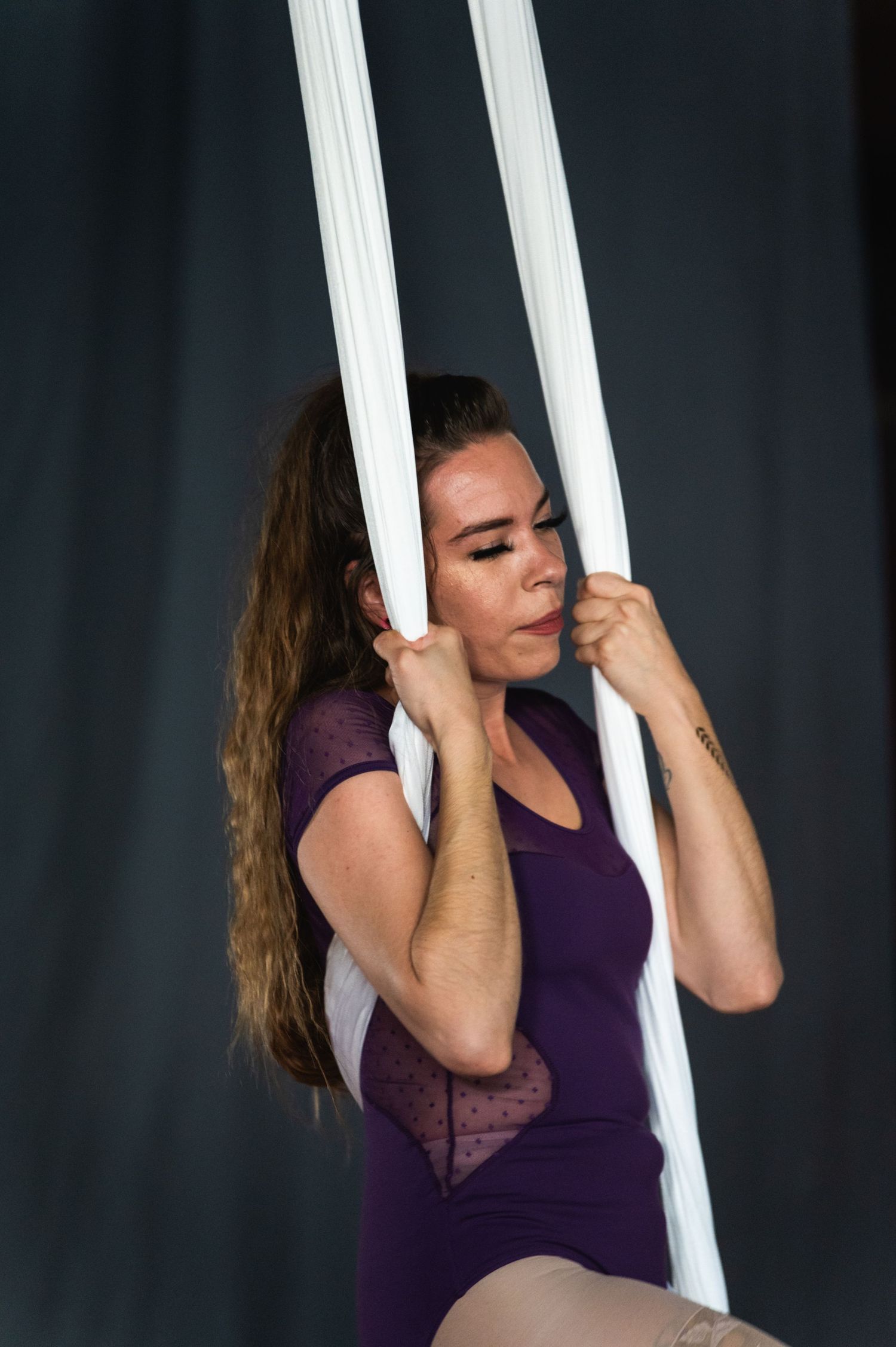 Aerial performer in purple leotard holds white silk straps during training session.