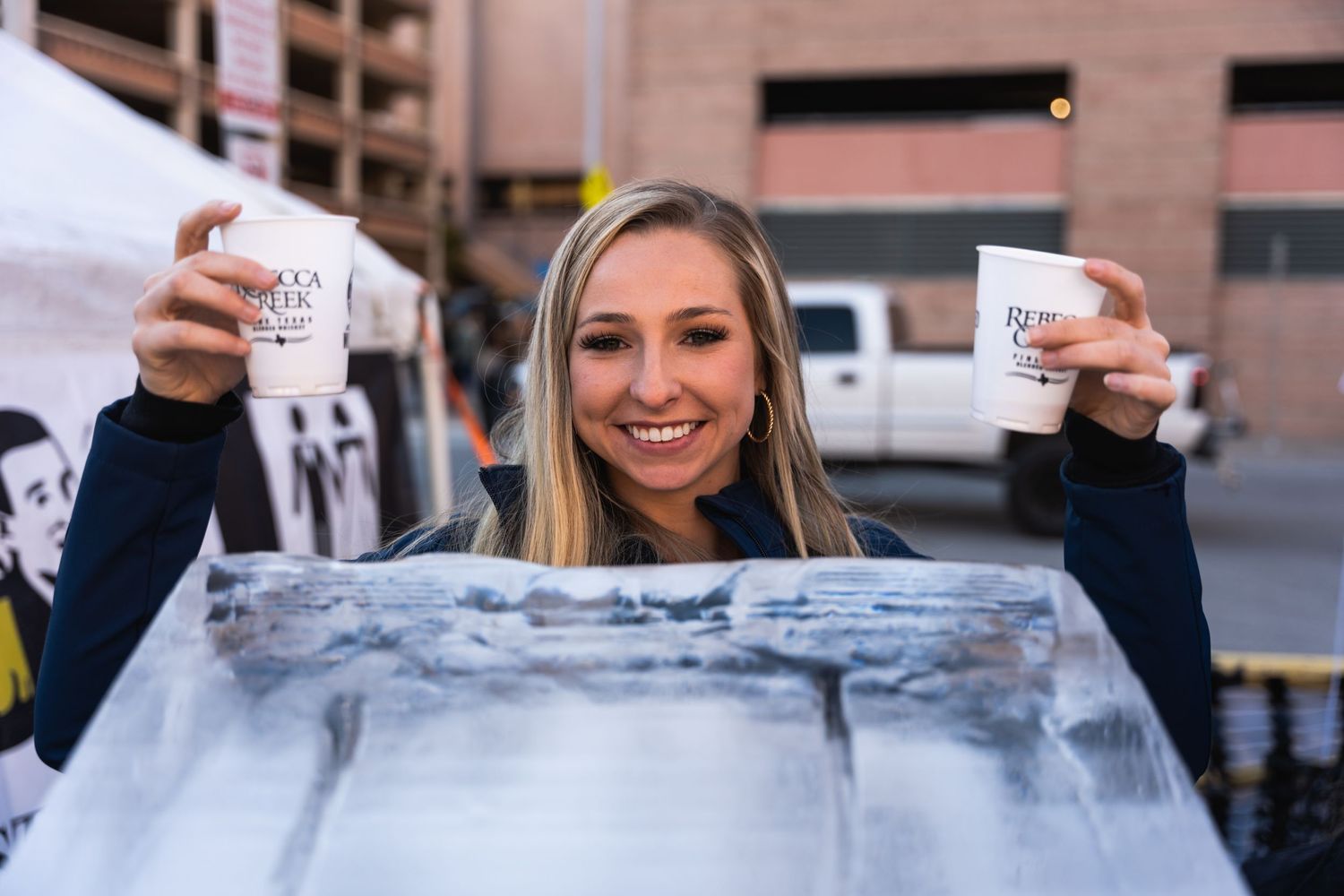 A person holds up two cups while posing with an ice luge at an outdoor event.