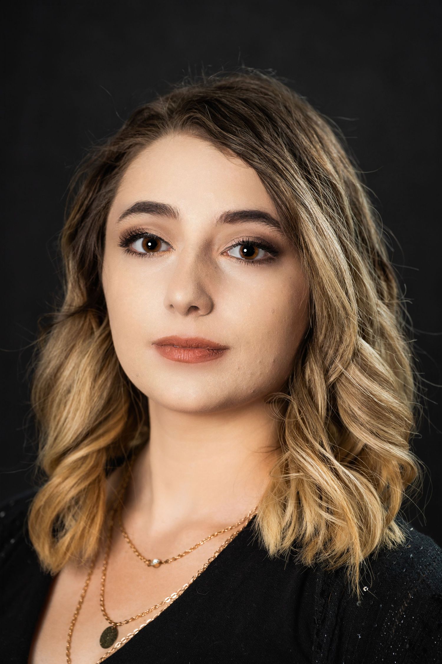 Professional portrait with wavy blonde hair and layered necklaces against dark background.