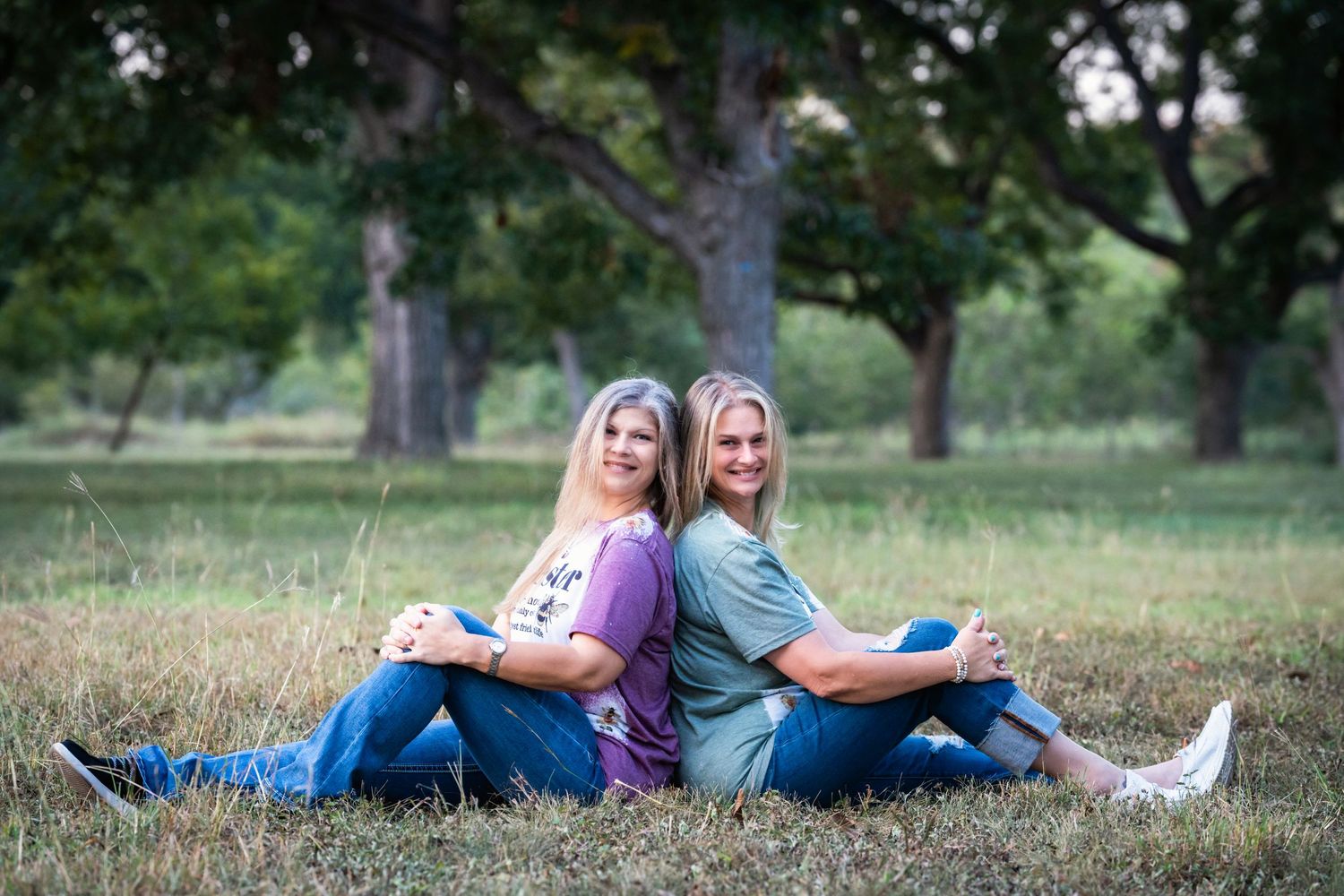 Two people in purple and teal shirts sit back-to-back on grass with trees in the background.
