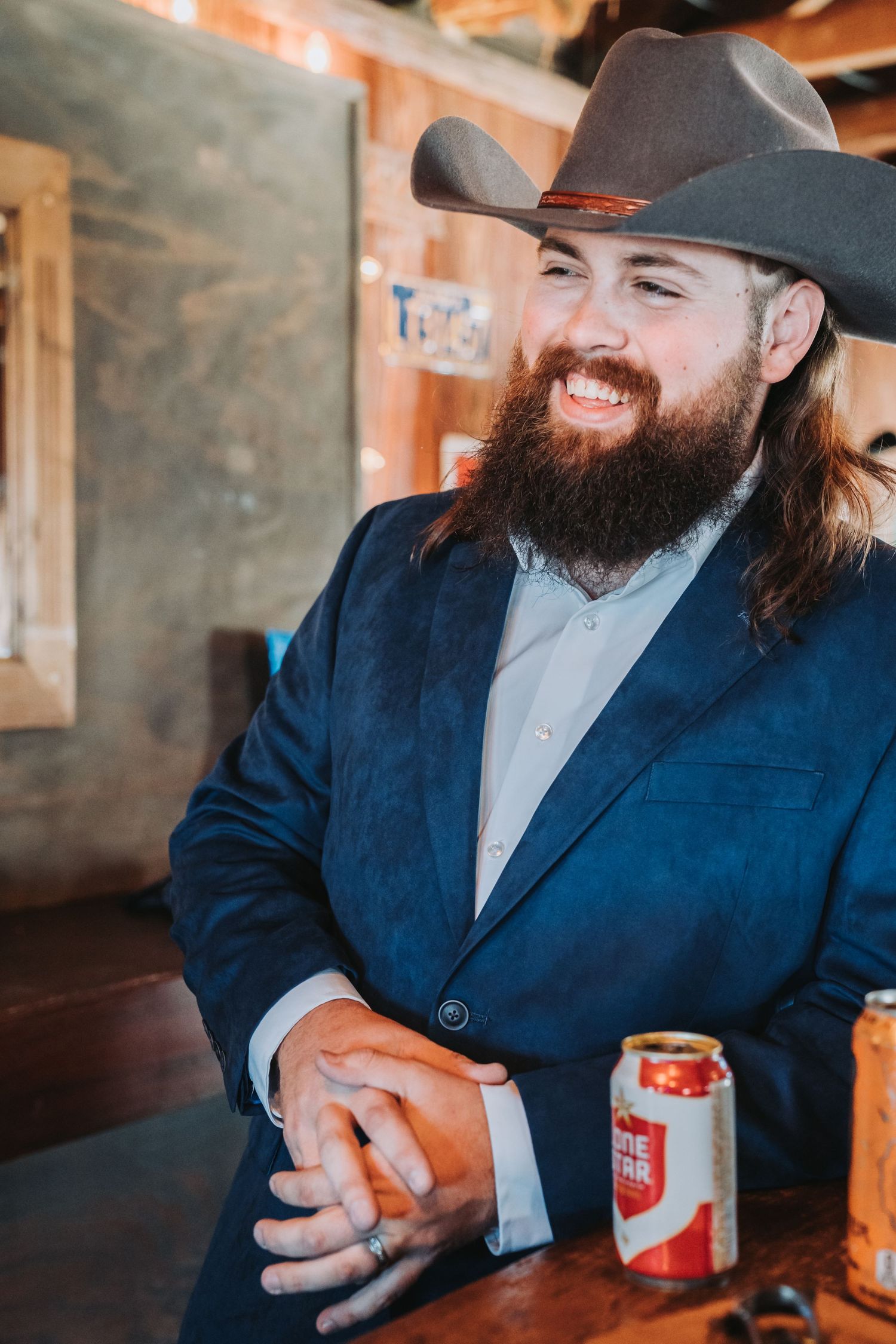 A bearded man in western attire smiles while sitting at a bar with a beverage.