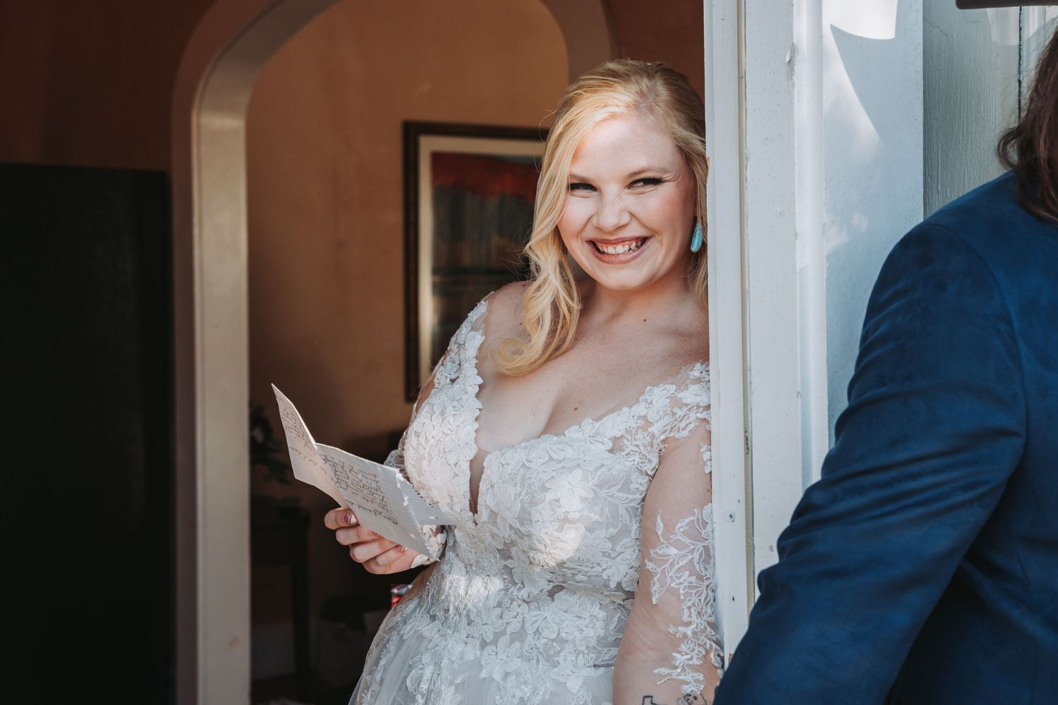 A person in a lace wedding dress holds a paper while standing in a doorway.