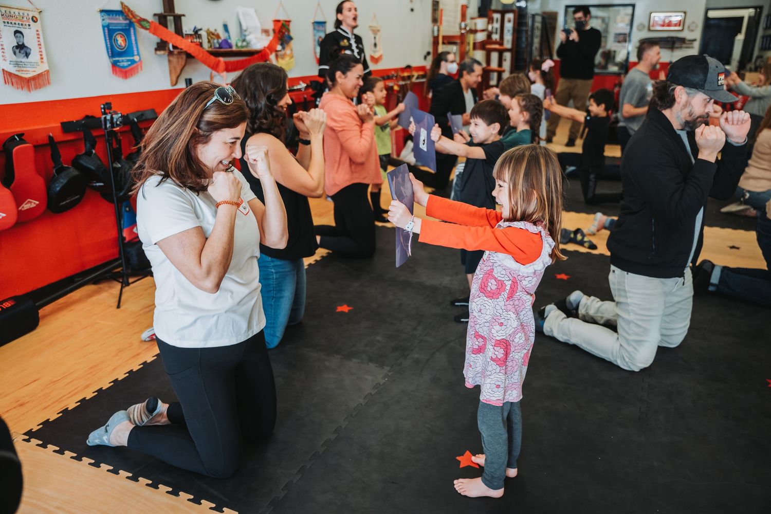 Parents and children practice martial arts moves together on padded floor mats.