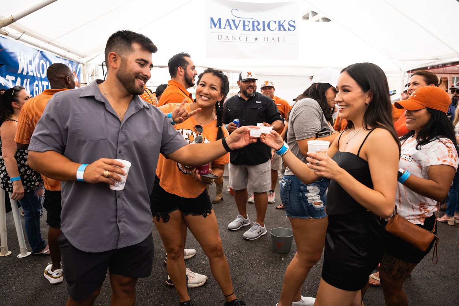 Party attendees in summer attire socializing at a branded outdoor event under a white tent.
