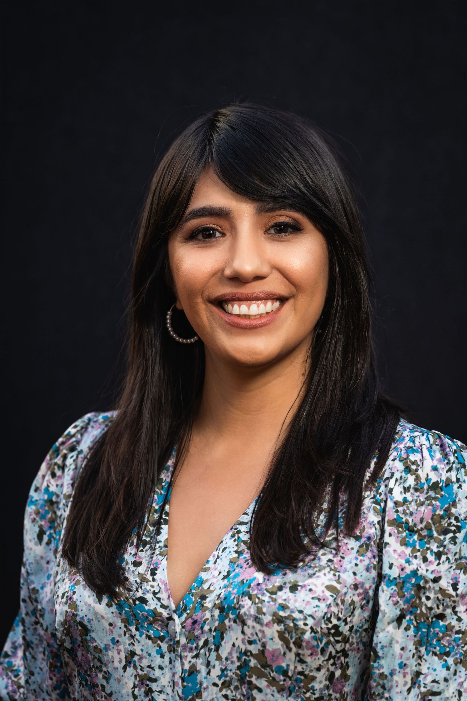 Professional portrait against dark background showing person in floral print top with silver hoop earrings.