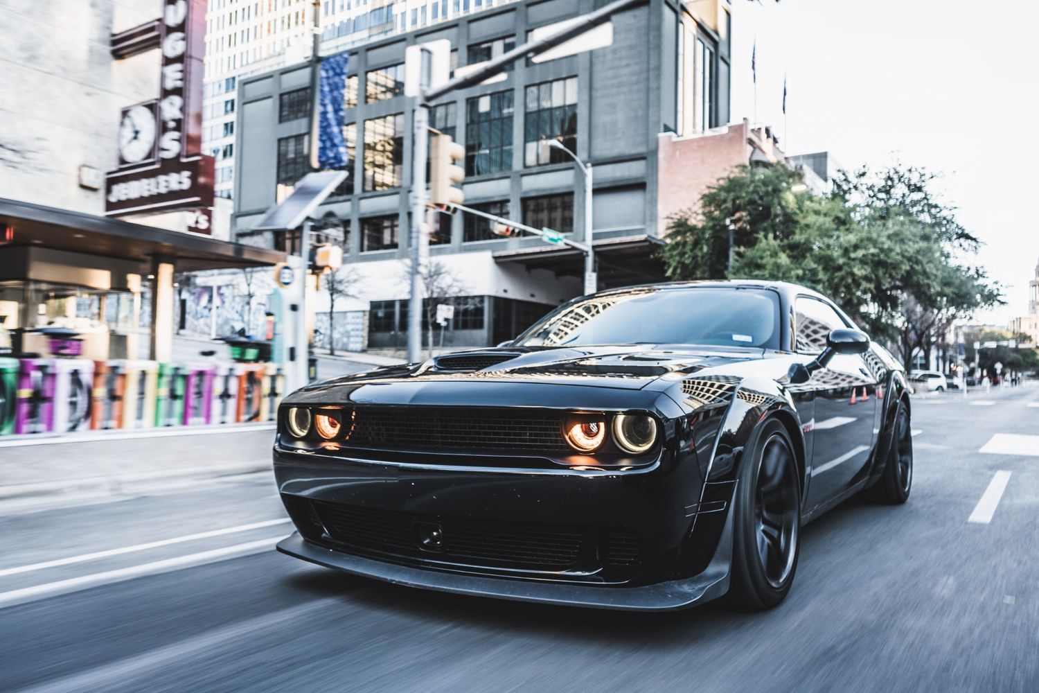 Black Dodge Challenger with custom headlights cruising through downtown at dusk.