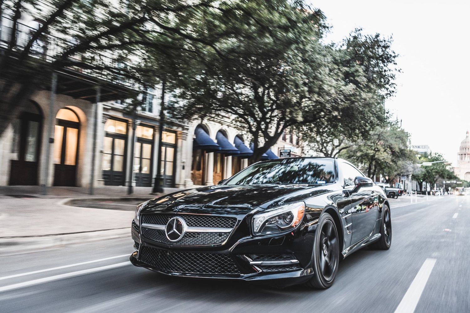 Black Mercedes-Benz sports car driving on tree-lined urban street.