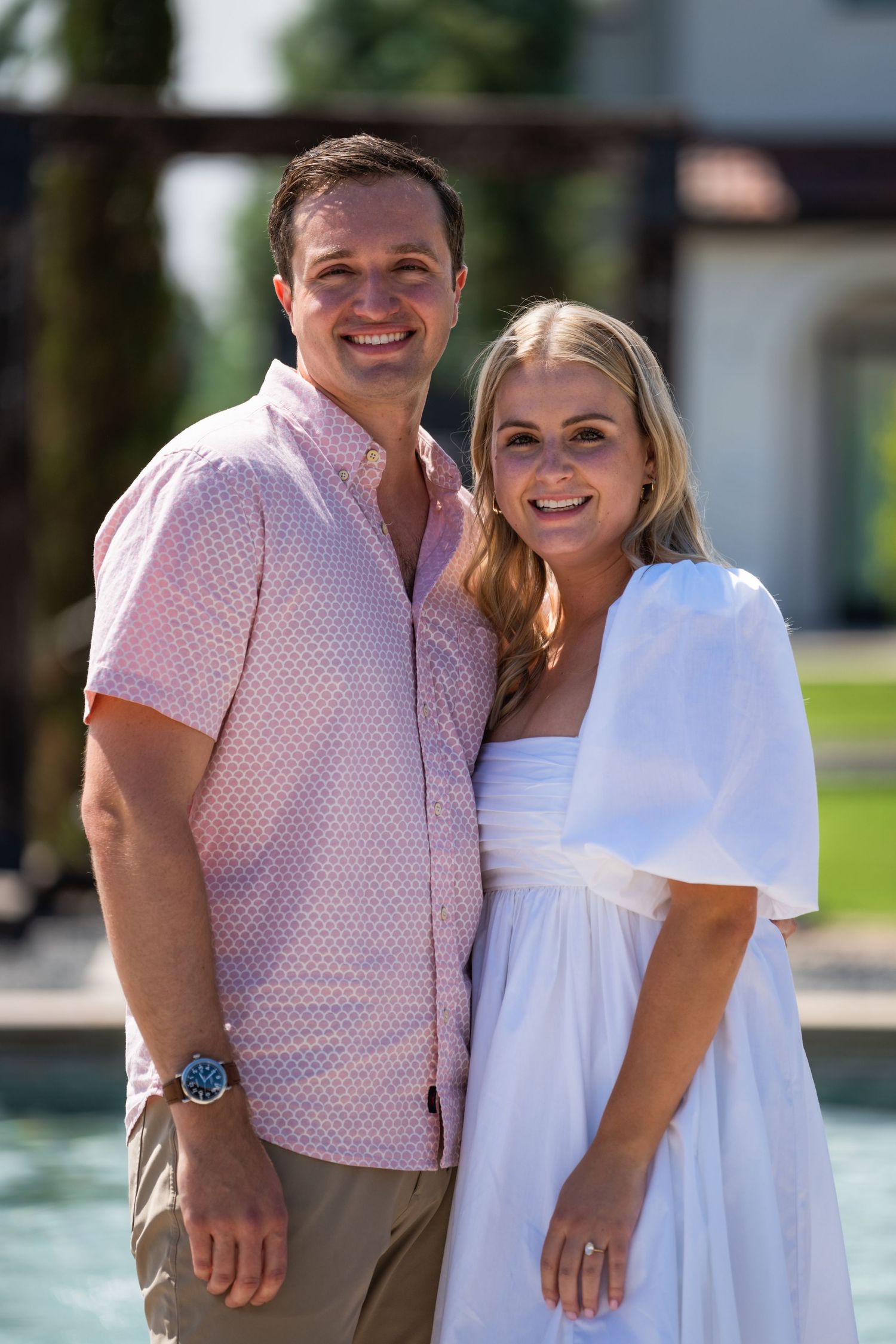 A couple in coordinating pink and white outfits smiles together by a pool at an elegant venue.