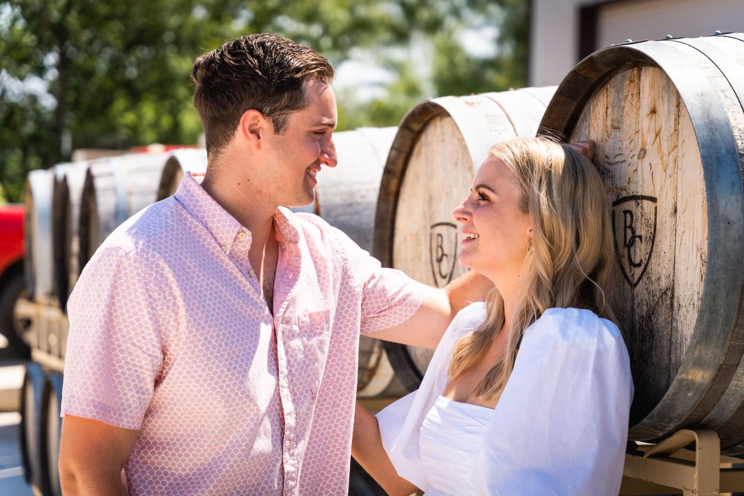 A couple shares a loving moment while standing against wooden wine barrels outdoors.