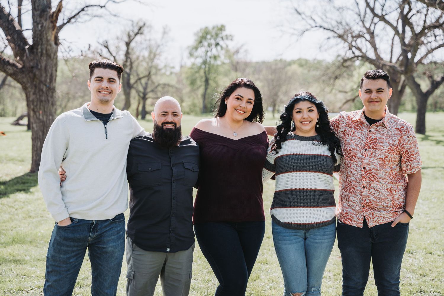Five people standing together in casual attire against a sunny spring landscape background.