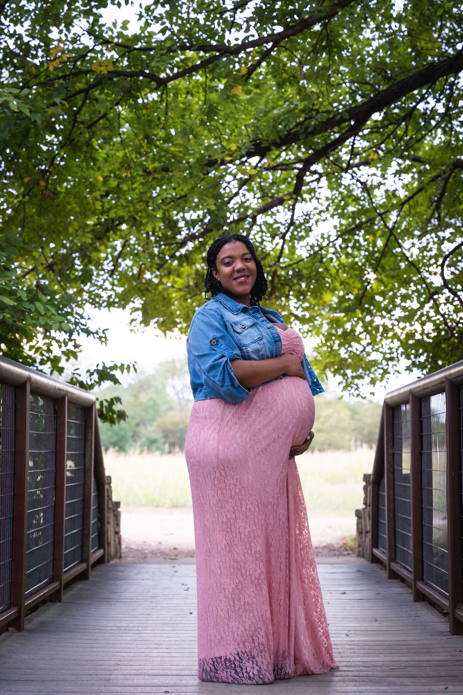 An expectant mother in a pink lace dress poses on a wooden bridge surrounded by green trees.