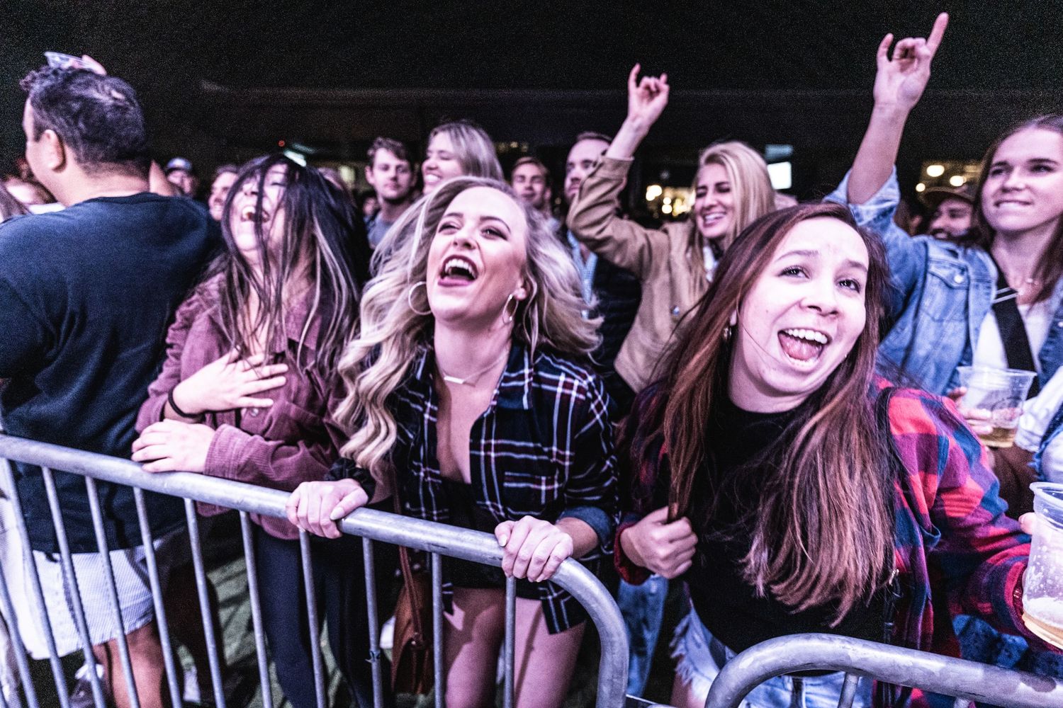 Excited concert attendees cheer and smile behind metal barricades at an outdoor music event.