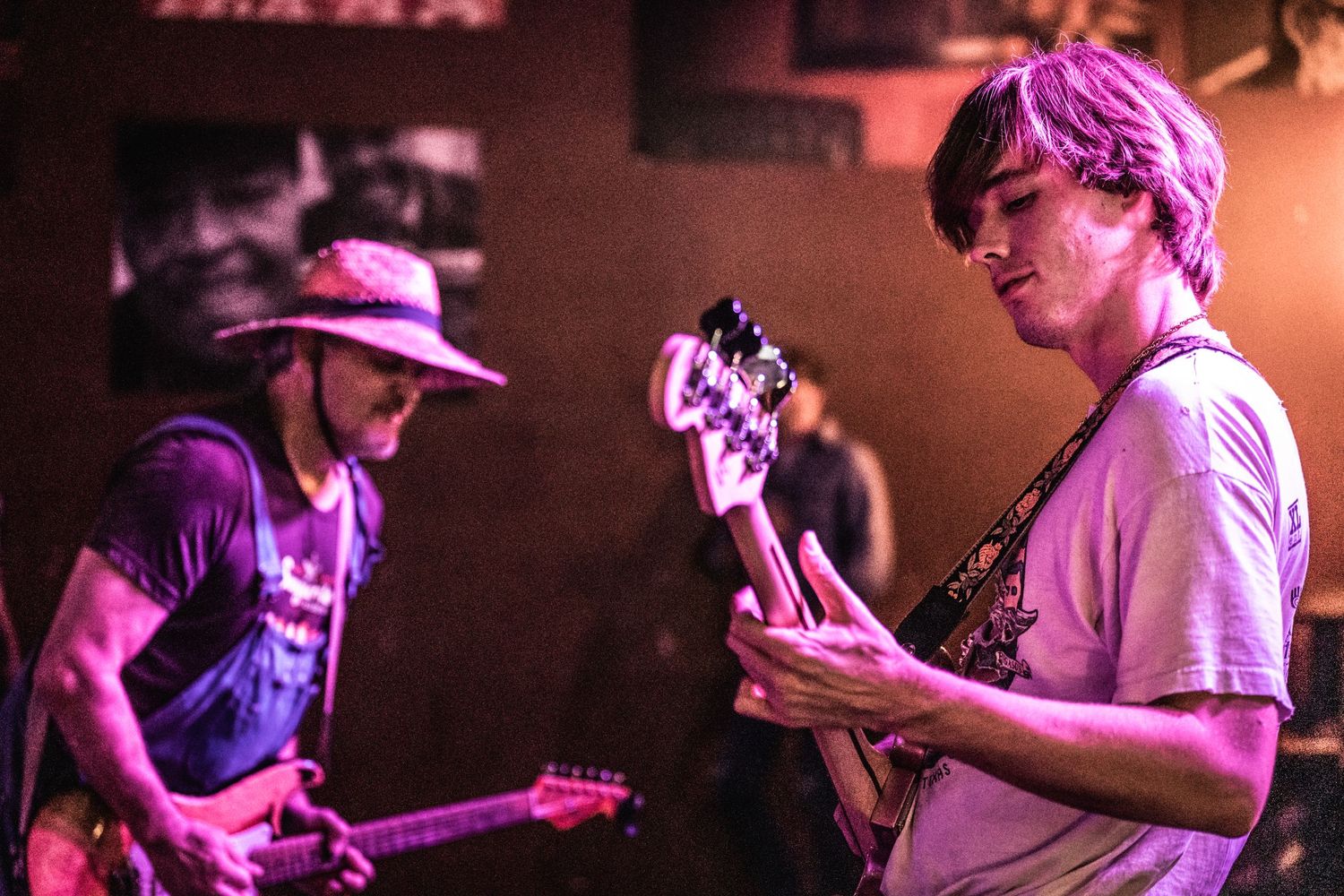 Two musicians playing guitars together during a live performance with purple-tinted lighting.
