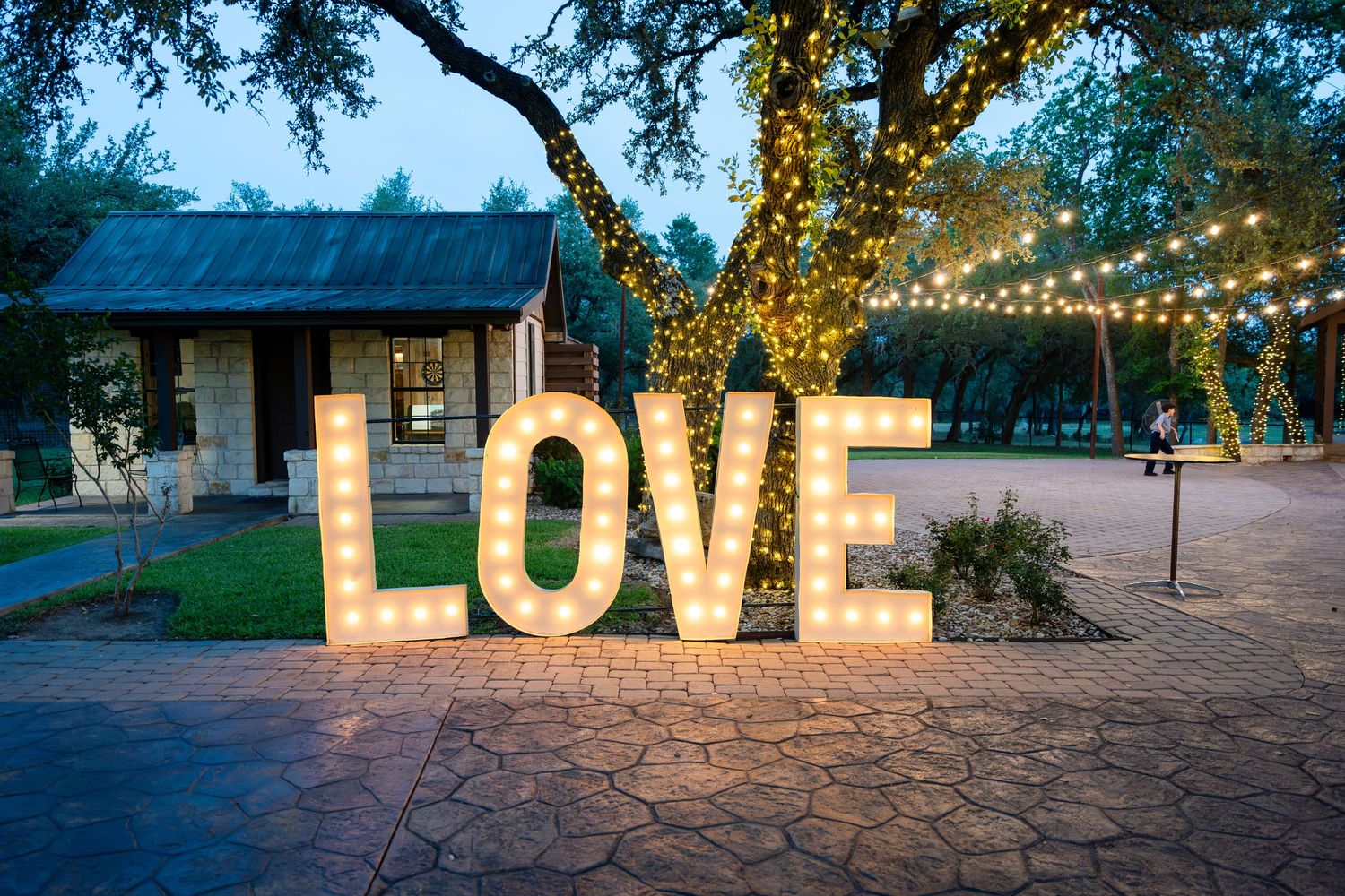 Large illuminated LOVE letters glow at dusk against rustic outdoor venue with string lights and stone patio.