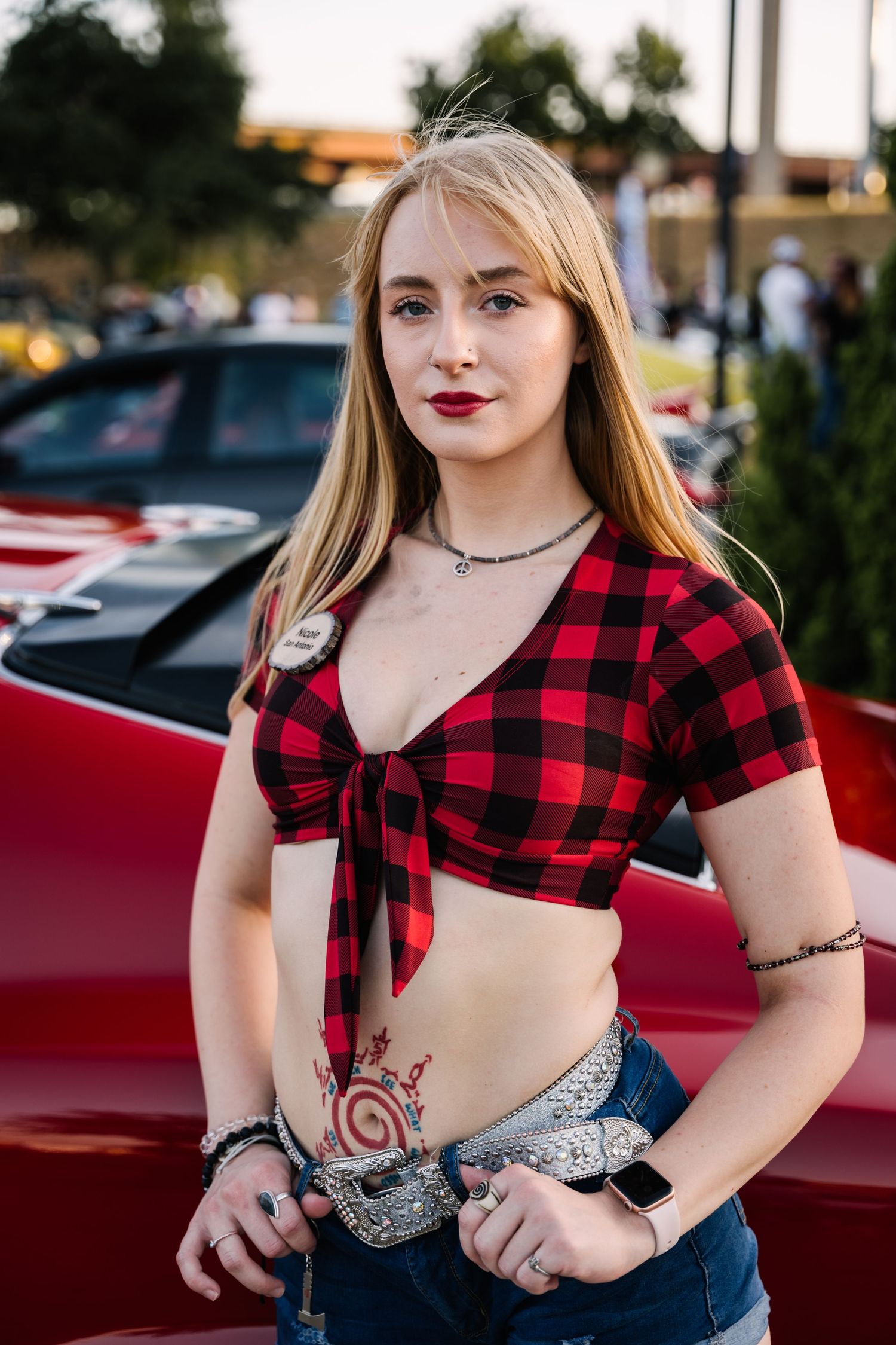 Person in red plaid tie-front top with denim shorts poses by classic car.