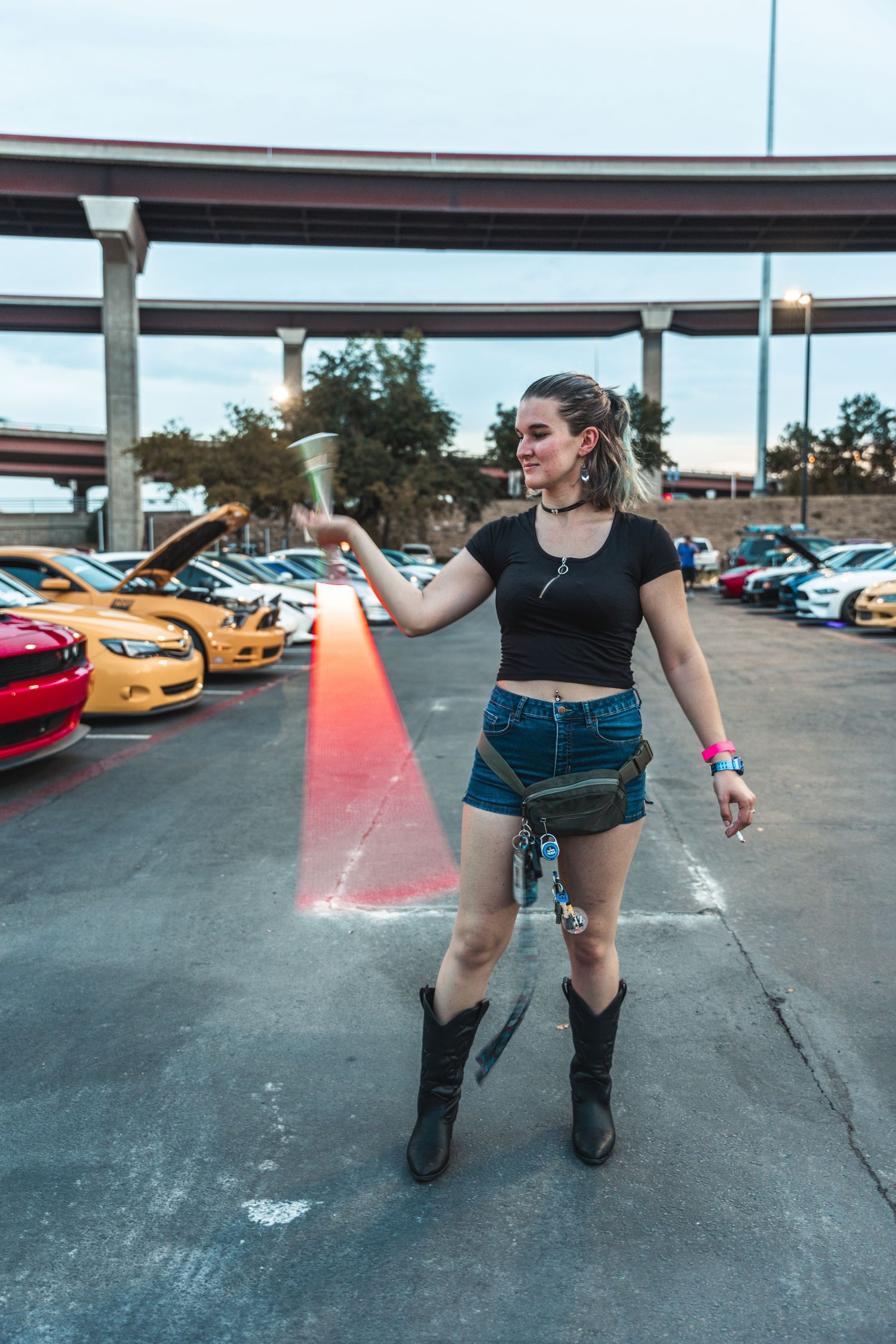 Person in a casual outfit holding a drink at a car meet parking lot.