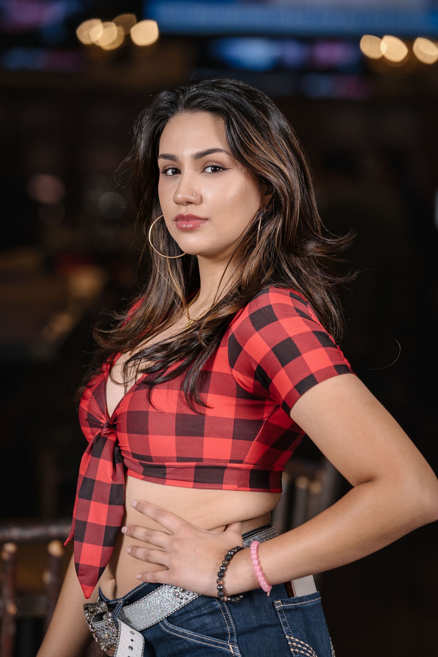 Model in red and black buffalo plaid crop top poses against blurred background with bokeh lights.
